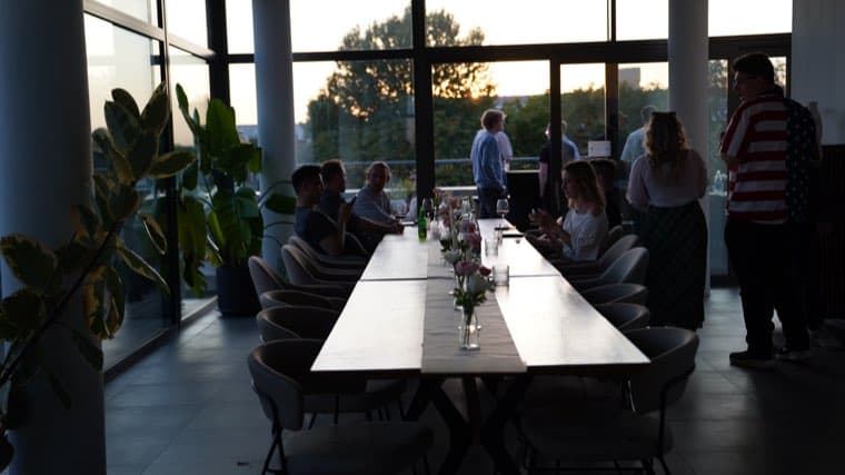 The long table at dusk, colleagues seated along it, the sun going down behind the terrace