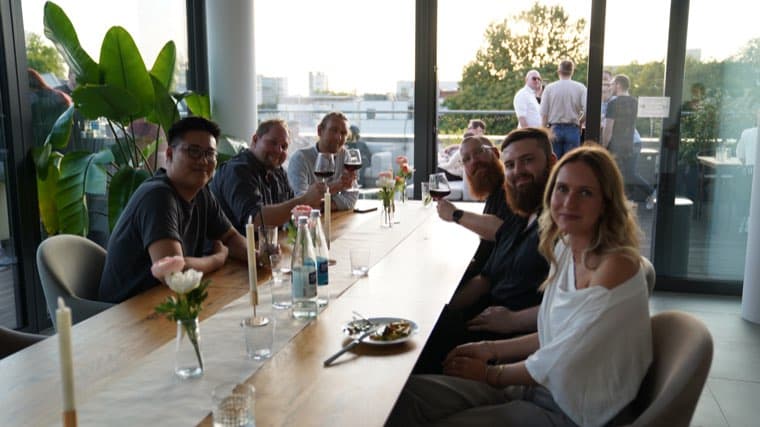 Seven colleagues around the long table in the office, turning to the camera during the company summer party