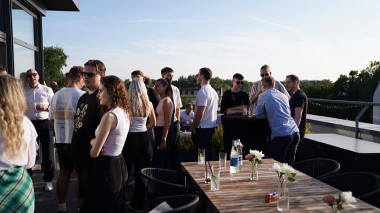 The roof terrace filling up: colleagues standing in groups with drinks under a clear sky