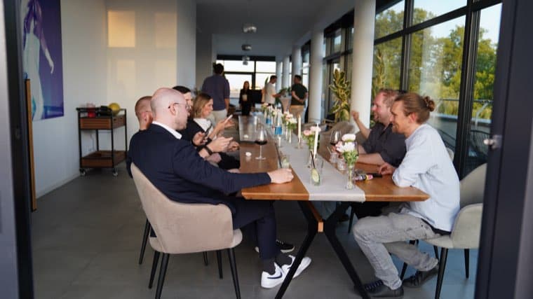 Colleagues talking across the long table in the office, the roof terrace visible through the glass behind them