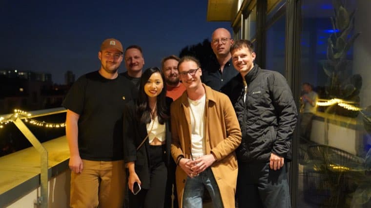 Seven colleagues on the terrace after dark, fairy lights strung along the railing behind them
