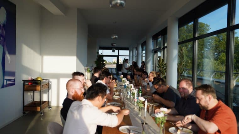 The full length of the long table in the office, around fifteen colleagues eating in the afternoon light from the window wall
