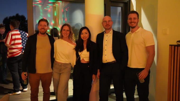 Five colleagues indoors late in the evening, coloured party lights on the wall behind them
